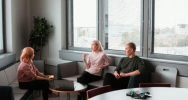 Three people are sitting and talking in a modern office lounge with large windows and a small table.