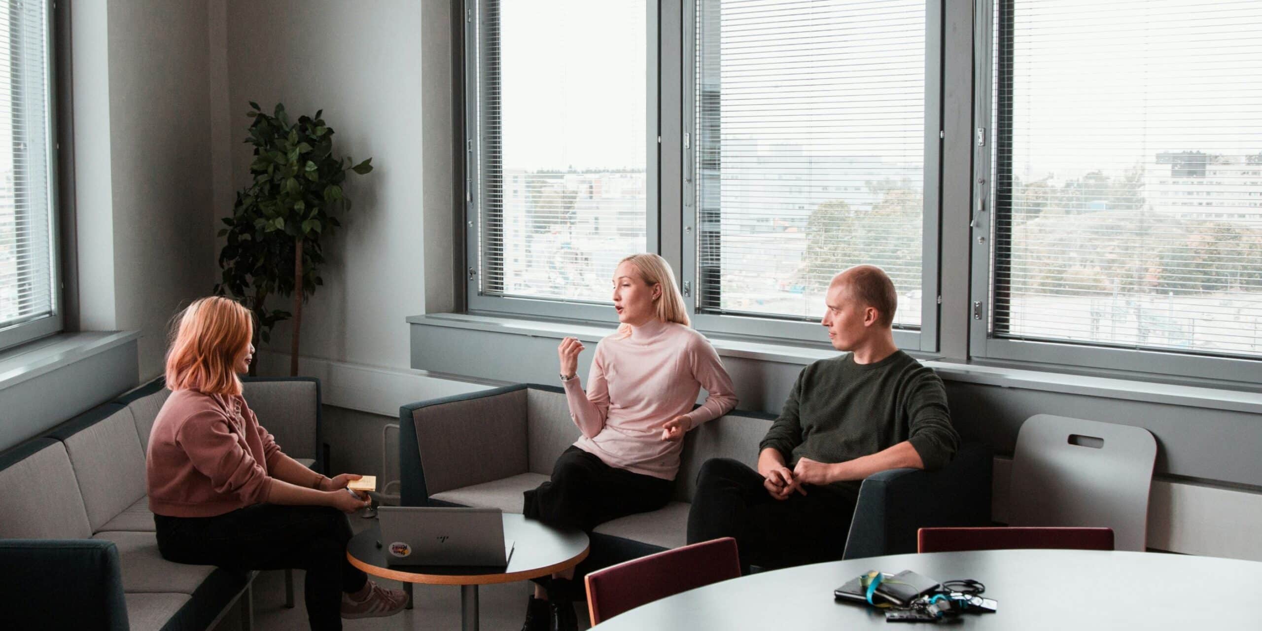 Three people are sitting and talking in a modern office lounge with large windows and a small table.