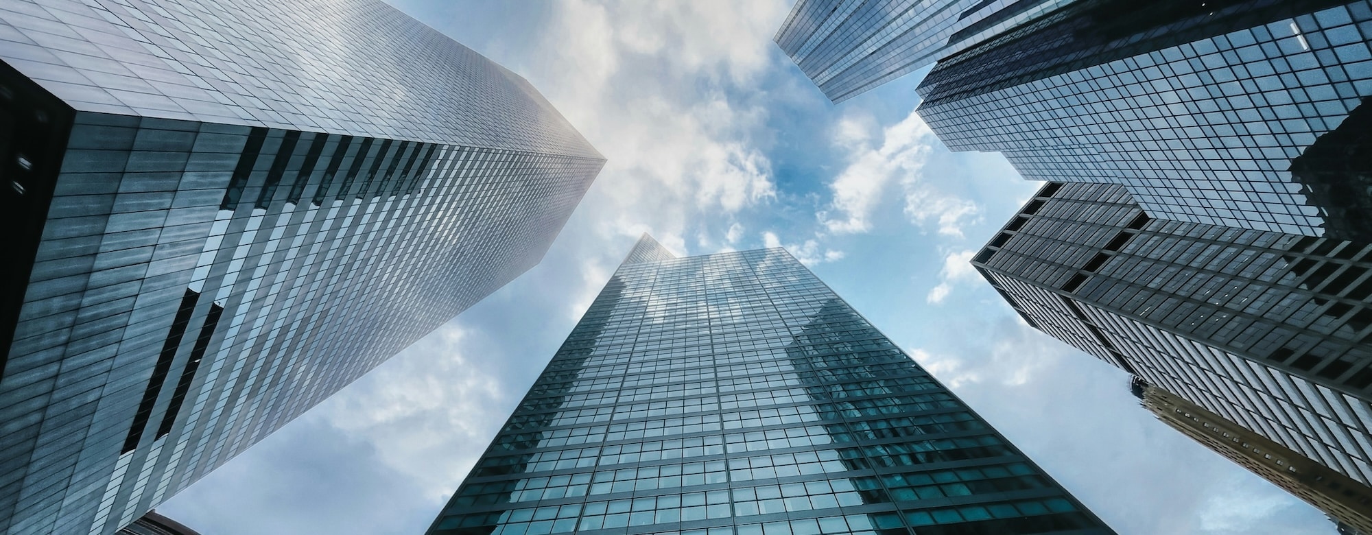 Looking up at four modern skyscrapers converging against a cloudy blue sky, creating a dramatic and symmetrical architectural perspective.