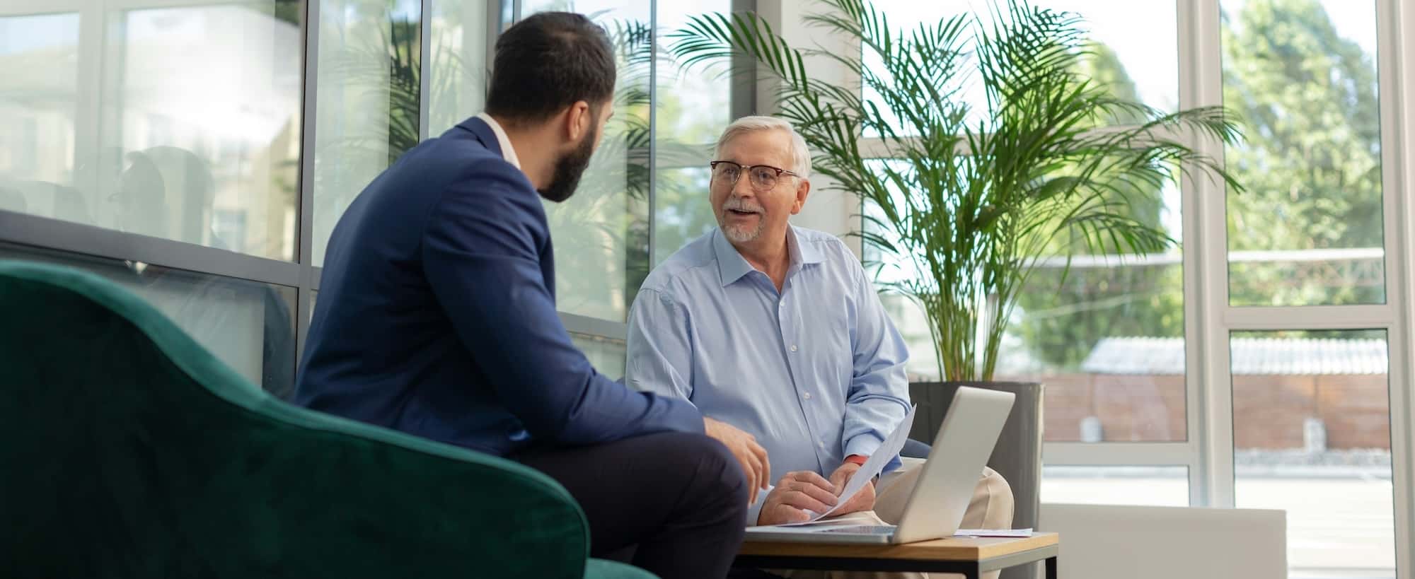 Two men are engaged in conversation in a bright, modern office setting with a laptop on the table and potted plants in the background.