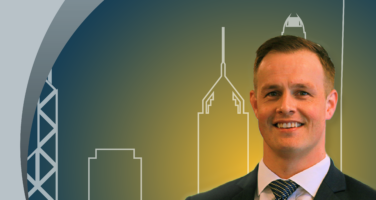 A man in a suit stands smiling against a backdrop of stylized city skyline graphics with his name written in the corner.