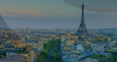 Aerial view of Paris with the Eiffel Tower prominently in the background, surrounded by historic buildings and tree-lined streets under a cloudy sky.