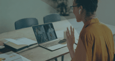 A person wearing earbuds is sitting at a table, participating in a video conference on a laptop, with documents spread around.
