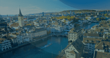 Aerial view of a historic European city with a river running through, featuring old buildings with brown roofs and a prominent church tower on a sunny day.