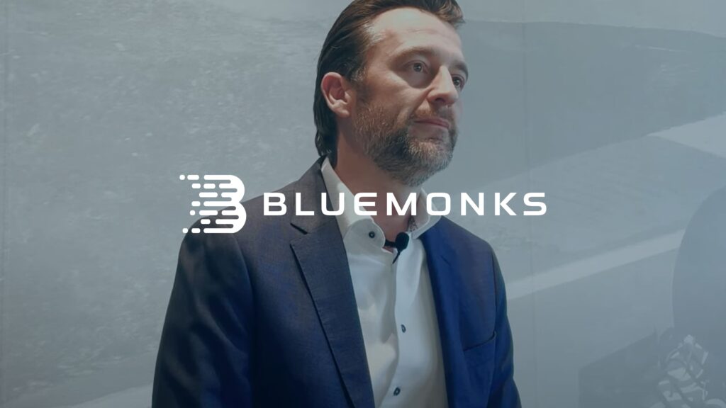 A man in a dark suit with a contemplative expression stands against a minimalist background. The word "BLUEMONKS" is prominently displayed over the image.