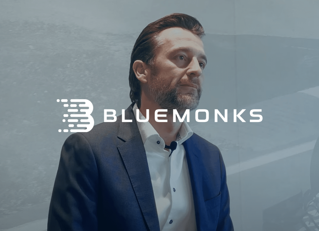 Man in a suit standing indoors with "Bluemonks" logo overlay.