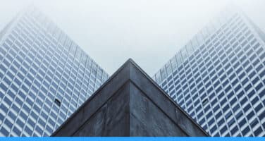 High-angle view of two modern skyscrapers with glass facades, converging at a sharp angle under a cloudy sky.