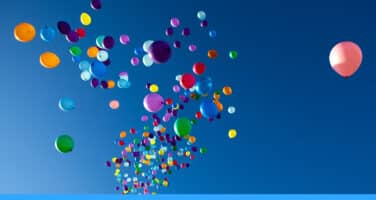 Numerous colorful balloons float upwards against a clear blue sky.
