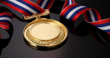 A gold medal with a laurel design and a red, white, and blue striped ribbon, placed on a reflective black surface.