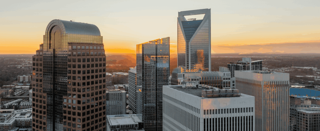 A city skyline at sunset, featuring tall modern skyscrapers with orange and pink hues in the sky.