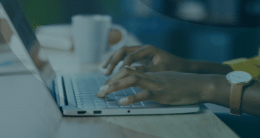 Person typing on a laptop with a white mug in the background, wearing a watch, and sitting at a wooden desk.