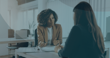 Two women are seated at a desk in a brightly lit office, engaged in a conversation. One woman gestures with her hands, while the other listens attentively.