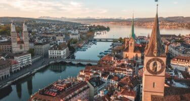 Aerial view of Zurich, showcasing historic buildings, two prominent church towers, and a river with bridges, set against a backdrop of distant mountains and a clear sky.