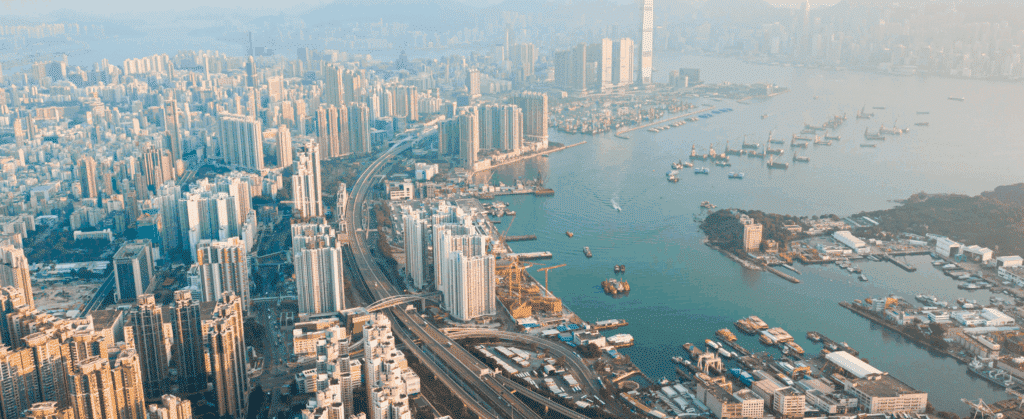 Aerial view of a bustling coastal city with numerous skyscrapers, a winding highway, and a harbor dotted with boats.