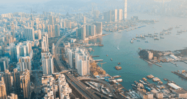 Aerial view of a bustling coastal city with numerous skyscrapers, a winding highway, and a harbor dotted with boats.