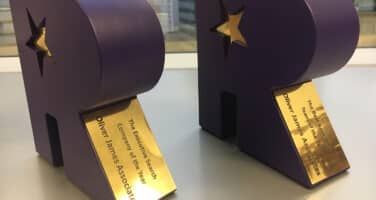 Two purple awards shaped like the letter R with gold star cutouts and plaques, placed on a table.