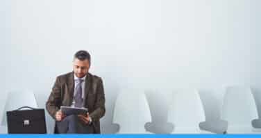 A man in a suit sits on a row of white chairs against a plain wall, holding a tablet with a briefcase beside him.