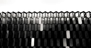 A black and white image of a modern building facade featuring a repetitive pattern of hexagonal glass panels, creating a geometric, abstract design against a bright sky.