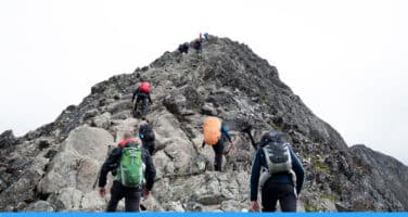 Hikers with backpacks climb a rocky mountain ridge under an overcast sky.