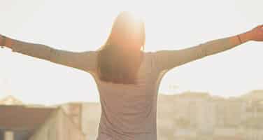 A person stands with outstretched arms, facing a bright sunlit cityscape.