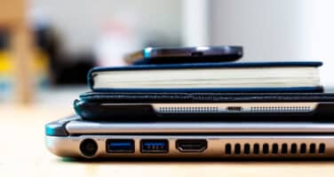 Stacked electronic devices and a notebook on a wooden surface, including a smartphone, a slim tablet, and an open laptop, showcasing their ports and connectors.