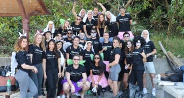 A group of people wearing matching black shirts and costumes gather outdoors, smiling and posing for a photo. They are standing on a paved area with trees and a covered bench in the background.