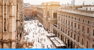 Aerial view of a bustling city square with historic architecture, including a prominent cathedral and ornate buildings. Crowds of people are scattered across the sunlit plaza, highlighting a vibrant urban scene.