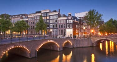 Illuminated canal bridges and traditional buildings reflect in the water at dusk, creating a serene urban scene.