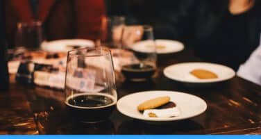 Glasses of dark beer and plates with cookies on a wooden table, surrounded by people seated in a cozy setting.