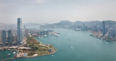 Aerial view of a bustling harbor with modern skyscrapers lining the coastline, surrounded by a mountainous backdrop under a hazy sky.