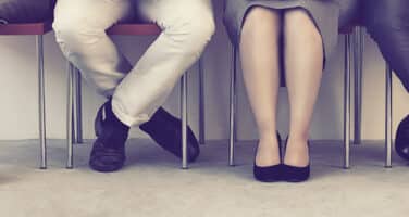 Four people seated on chairs in a row, showing only their legs and crossed ankles.