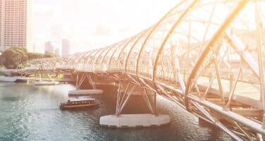 A modern, curved pedestrian bridge with intricate metal design stretches over a calm river, with a boat nearby and city buildings in the background under a bright sky.