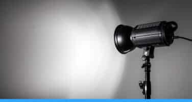 A studio spotlight on a stand casts a bright circle of light onto a plain wall, creating a stark contrast between light and shadow.