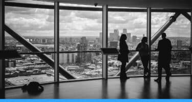 Three people stand in silhouette inside a high-rise building, looking out at a panoramic cityscape through large windows, with a cloudy sky in the background.