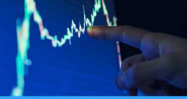 A hand pointing at a fluctuating line graph on a blue screen, indicating data analysis or stock market trends.