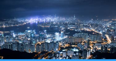 Aerial view of a sprawling cityscape at night, with illuminated skyscrapers and buildings densely packed together, surrounding a dark harbor.