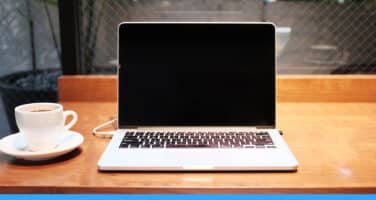 A laptop with a blank screen sits on a wooden table next to a cup of coffee, in a cozy indoor setting.