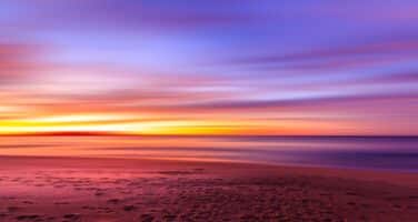 Vibrant sunset over a peaceful beach, with a gradient sky transitioning from purple to orange, and blurred ocean waves meeting the smooth sandy shore.