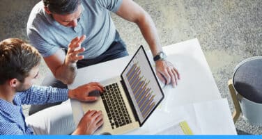 Two people engage in discussion over a laptop displaying a chart, seated at a white table in a modern office setting.