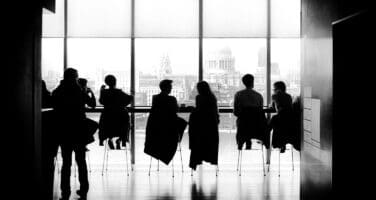 Silhouetted group of people seated at a high counter, facing large windows with an urban skyline in the background.