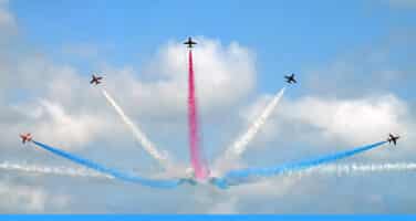 Five aerobatic planes create a fan-shaped formation, releasing trails of red, white, and blue smoke against a blue sky with soft clouds.