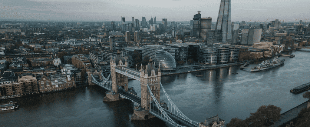 Aerial view of Tower Bridge spanning the River Thames in London, with modern skyscrapers and historic buildings in the background under a cloudy sky.