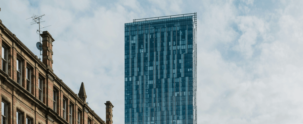 A modern glass skyscraper rises against a cloudy sky, with part of an older brick building visible in the foreground.