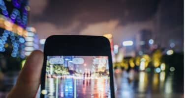 A person holding a smartphone captures a vibrant, colorful cityscape at night, with buildings and lights reflecting on a wet surface creating a blurred effect.