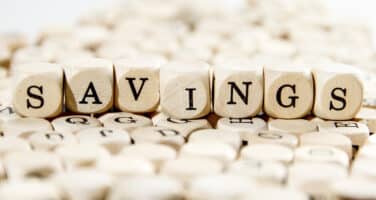 Wooden cubes spelling "SAVINGS" against a blurred background of other cubes with letters.
