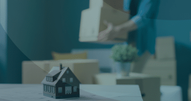 A small model house is in focus in the foreground, while in the background, a person carries moving boxes in a room filled with packed items.