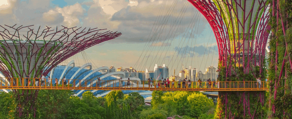 An elevated walkway connects large vertical gardens with colorful trunks, set against a city skyline and lush greenery under a partly cloudy sky.