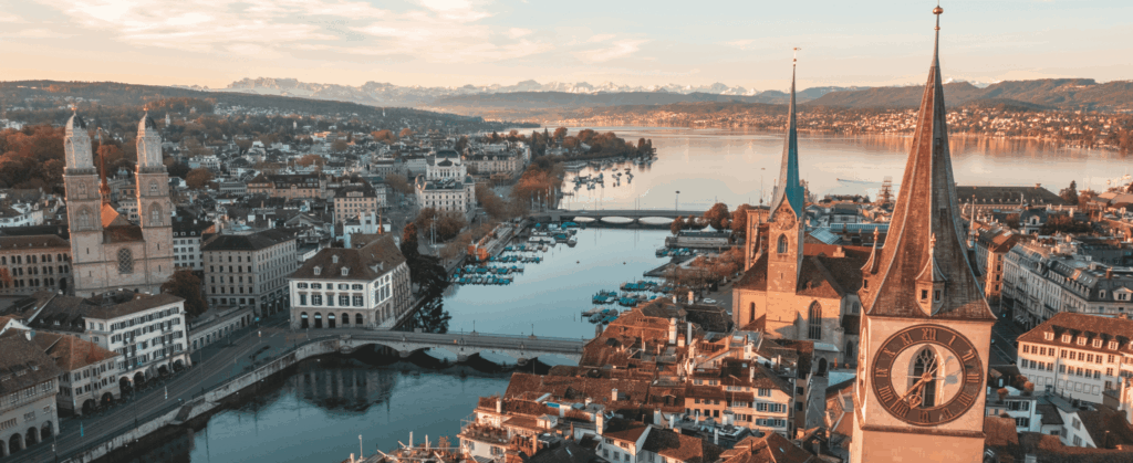 Aerial view of a historic European city with a river, prominent clock tower, and surrounding buildings, set against a backdrop of distant mountains under a partly cloudy sky.