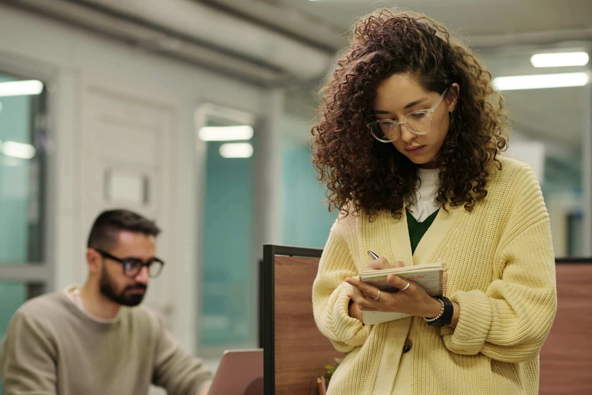 A woman with curly hair writes in a notebook while standing. In the background, a man with glasses works on a laptop at a desk in an office setting.