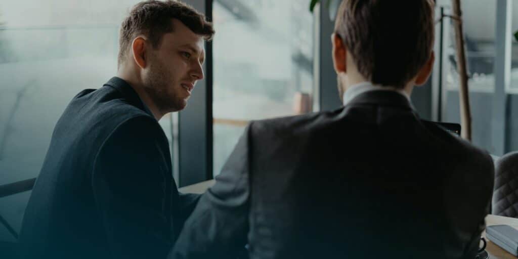Two men in suits are engaged in conversation at a modern office with large windows, suggesting a professional setting.